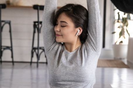 Home Fitness - Cheerful young ethnic female in earbuds sitting on yoga mat with crossed legs and closed eyes while listening to music and raising hands in apartment