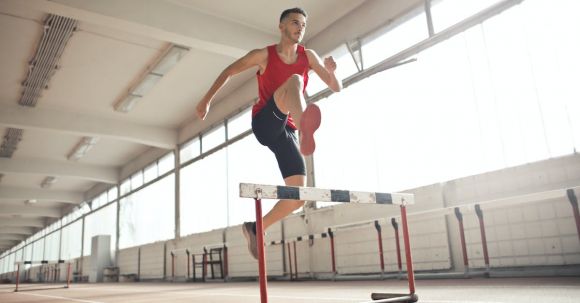 Speed Training - Powerful male athlete jumping over hurdle on running track while training at sports hall