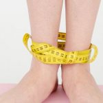 Weight Loss Programs - High angle of crop anonymous female with centimeter on legs checking weight on scales on white background