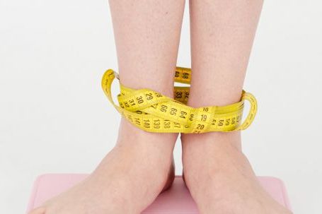 Weight Loss Programs - High angle of crop anonymous female with centimeter on legs checking weight on scales on white background