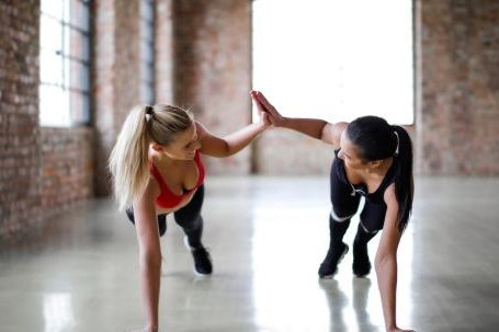 Fitness Support - Fit smiling women in sportswear looking at each other and giving high five while doing push up exercise on gray glossy floor against blurred interior of spacious workout room with brick walls and big windows