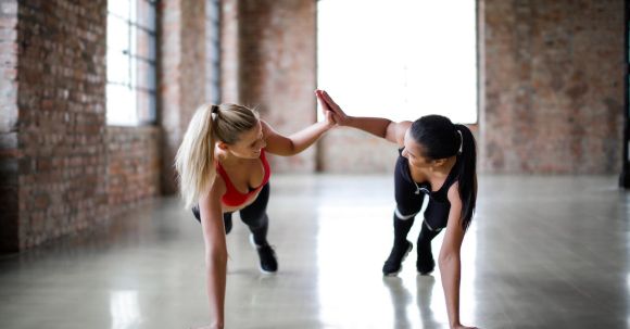 Fitness Support - Fit smiling women in sportswear looking at each other and giving high five while doing push up exercise on gray glossy floor against blurred interior of spacious workout room with brick walls and big windows