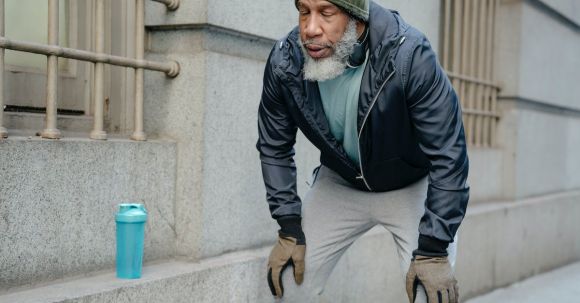 Lean Strength - Concentrated bearded senior black man wearing warm sportswear leaning forward with hands on knees and closed eyes in street near building while break in workout