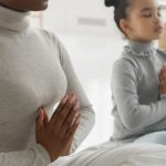 Yoga Integration - Full body of tranquil young barefooted African American woman in casual clothes sitting on carpet in Padmasana pose with closed eyes while meditating with adorable little daughter at home