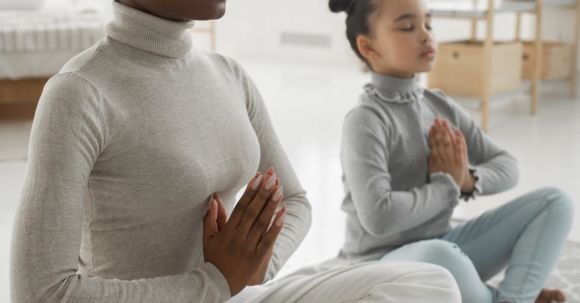 Yoga Integration - Full body of tranquil young barefooted African American woman in casual clothes sitting on carpet in Padmasana pose with closed eyes while meditating with adorable little daughter at home