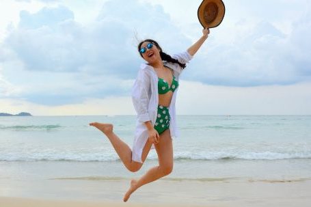 Long-term Success - Woman Jumping on Seashore and Holding Hat