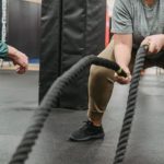 Fit Challenge - Strong African American female in activewear training with battle ropes near fit male coach in fitness center