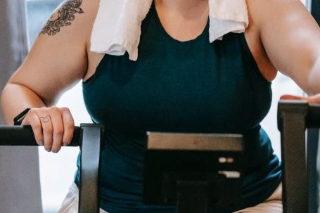 Fit Challenge - Smiling plus size female with towel exercising on cross trainer machine while looking at camera during workout in fitness studio