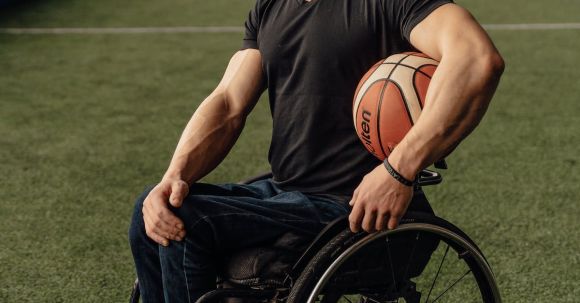 Sports Training - Man in Black Shirt and Blue Denim Jeans Sitting on Wheelchair Holding Basketball