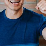 Fitness Success - Cheerful young ethnic male athlete in sportswear smiling and showing fist while standing against brick wall after successful workout in gym