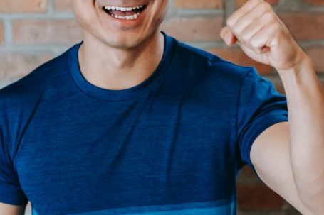 Fitness Success - Cheerful young ethnic male athlete in sportswear smiling and showing fist while standing against brick wall after successful workout in gym