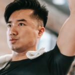 Healthy Challenge - Sportive Asian male in black sportswear with raised arm lifting metal dumbbell while training in fitness studio with woman on blurred background