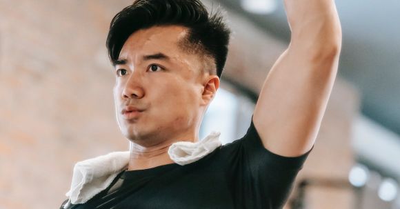 Healthy Challenge - Sportive Asian male in black sportswear with raised arm lifting metal dumbbell while training in fitness studio with woman on blurred background