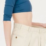 Fitness Gains - Side view of slim positive female with bare belly wearing oversized trousers standing on white background in light studio after weight loss