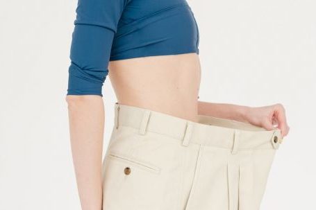 Fitness Gains - Side view of slim positive female with bare belly wearing oversized trousers standing on white background in light studio after weight loss