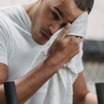 Pre-workout Fuel, Post-workout Recovery. - Young male wiping forehead with towel during rest after hard workout in contemporary fitness center
